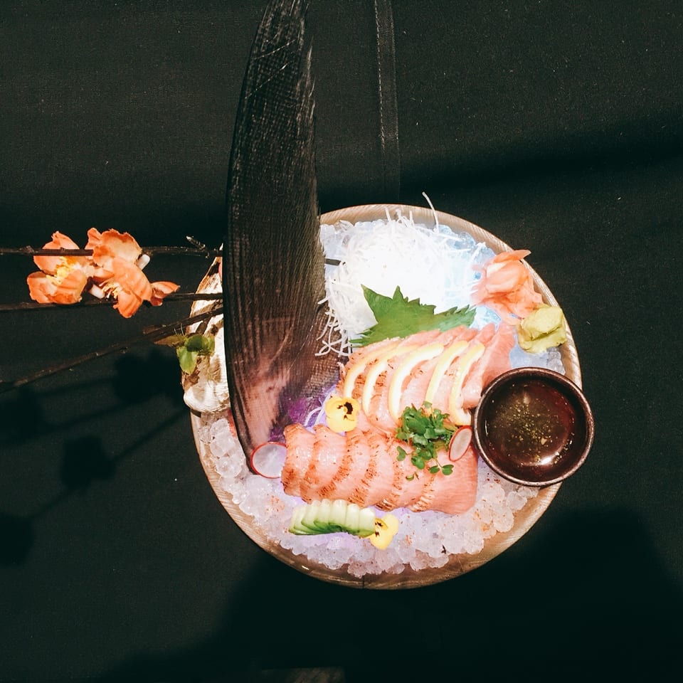 Sashimi platter with sliced fish, lemon, and garnish on ice, with a dark sauce and decorative flowers.
